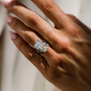 May include: A ring featuring a large, rectangular radiant-cut clear gemstone set on a thin, polished gold band. The stone has multiple facets that reflect light, creating a brilliant appearance. The ring is worn on a finger, showcasing the stone's size.