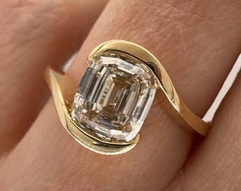 May include: A gold ring featuring a large, rectangular-cut diamond. The diamond is set in a bezel setting with a swirling gold band. The ring is shown on a person's finger, showcasing the design and sparkle of the gemstone.