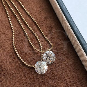 May include: Two gold necklaces with large, round, clear diamond pendants. The necklaces have delicate beaded chains. The pendants are set in gold and sparkle brightly. The jewelry is displayed on a brown surface, with a green and beige box in the background.
