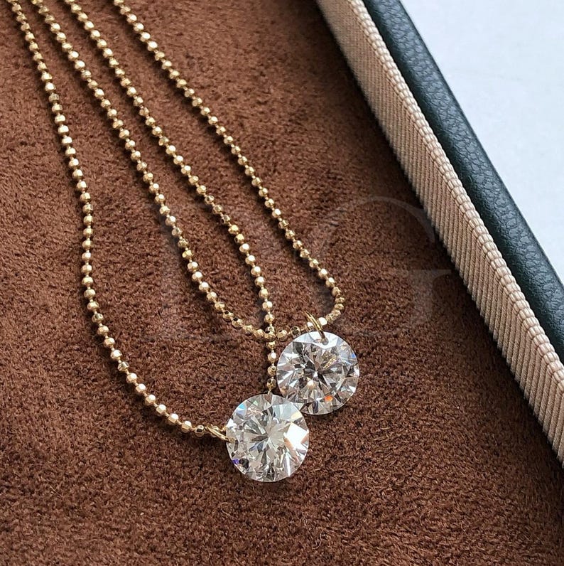May include: Two gold necklaces with large, round, clear diamond pendants. The necklaces have delicate beaded chains. The pendants are set in gold and sparkle brightly. The jewelry is displayed on a brown surface, with a green and beige box in the background.