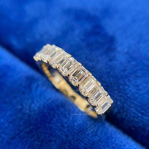 Puede incluir: Un anillo de banda de oro con una serie de piedras preciosas claras de corte rectangular. El anillo se muestra sobre un fondo azul vibrante, resaltando el brillo de las piedras. Una joya.