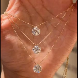May include: Three-strand gold necklace with round, clear gemstones. The necklaces are layered, with each strand featuring a single gemstone pendant. The jewelry is held in a hand, showcasing the delicate design and sparkle.