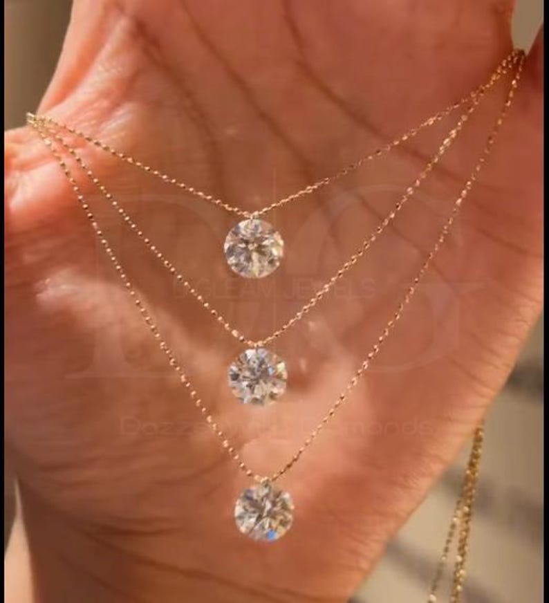 May include: Three-strand gold necklace with round, clear gemstones. The necklaces are layered, with each strand featuring a single gemstone pendant. The jewelry is held in a hand, showcasing the delicate design and sparkle.