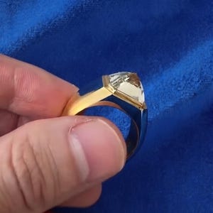 May include: A gold ring featuring a prominent, clear, triangular-cut gemstone set in a bezel setting. The band is wide and smooth, showcasing a polished metallic finish that reflects light. The design highlights the geometric shape of the stone.