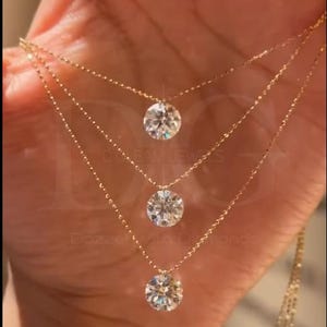 May include: Three-strand gold necklace featuring round, clear gemstones. The delicate chains are connected to the gemstones, creating a layered effect. The jewelry is displayed against a warm-toned background, highlighting the sparkle of the stones.