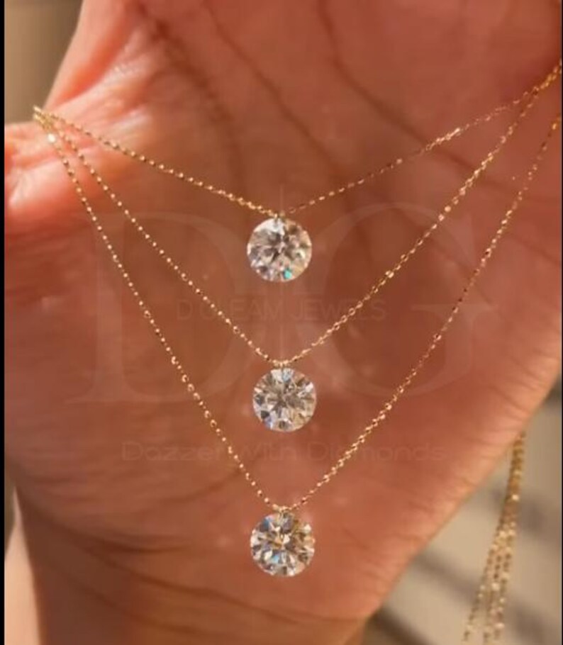 May include: Three-strand gold necklace featuring round, clear gemstones. The delicate chains are connected to the gemstones, creating a layered effect. The jewelry is displayed against a warm-toned background, highlighting the sparkle of the stones.