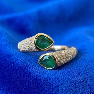 May include: A gold-toned ring with two pear-shaped, deep green gemstones. The ring's band is encrusted with small, clear gemstones. The ring is set against a vibrant blue background. The text "OOLEAM JEWELS" is visible on the ring.