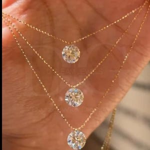 May include: Three delicate gold necklaces, each featuring a sparkling round diamond pendant. The chains are finely crafted, and the diamonds are set to maximize brilliance. The jewelry is displayed against a warm-toned background.