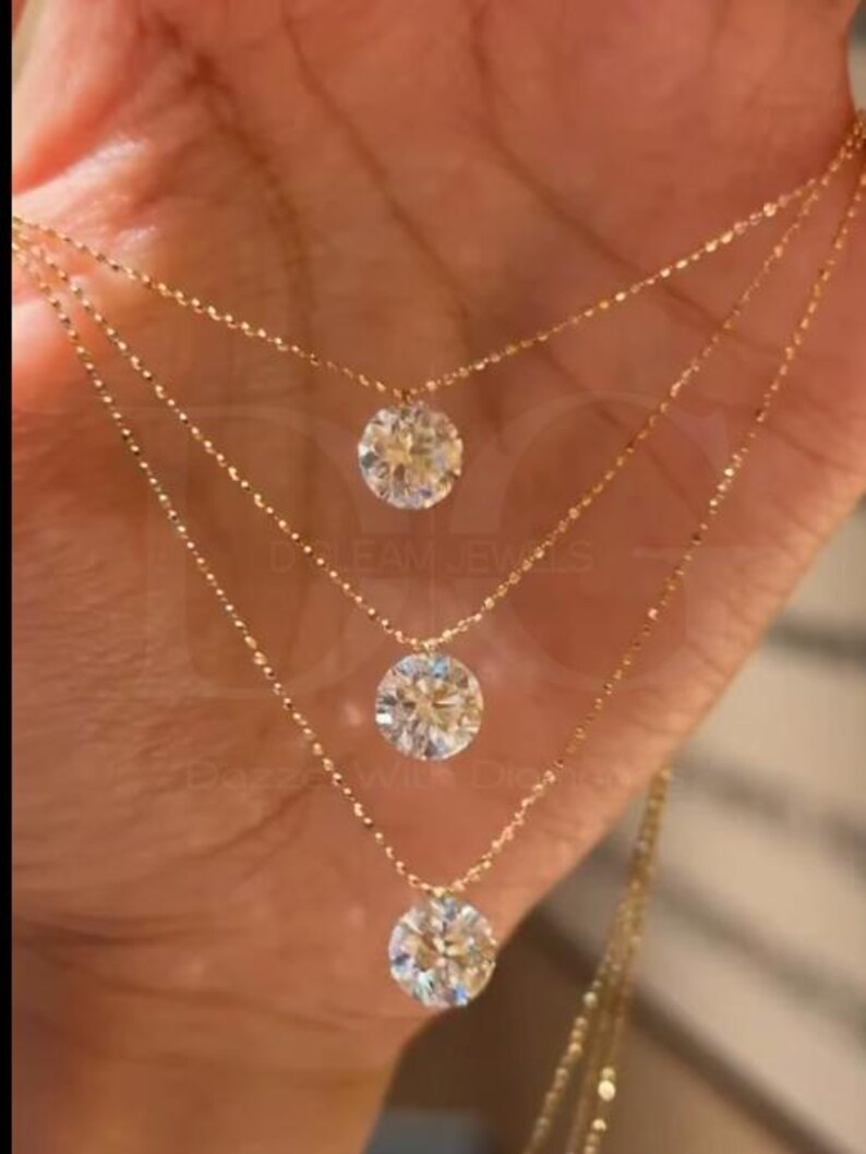 May include: Three delicate gold necklaces, each featuring a sparkling round diamond pendant. The chains are finely crafted, and the diamonds are set to maximize brilliance. The jewelry is displayed against a warm-toned background.