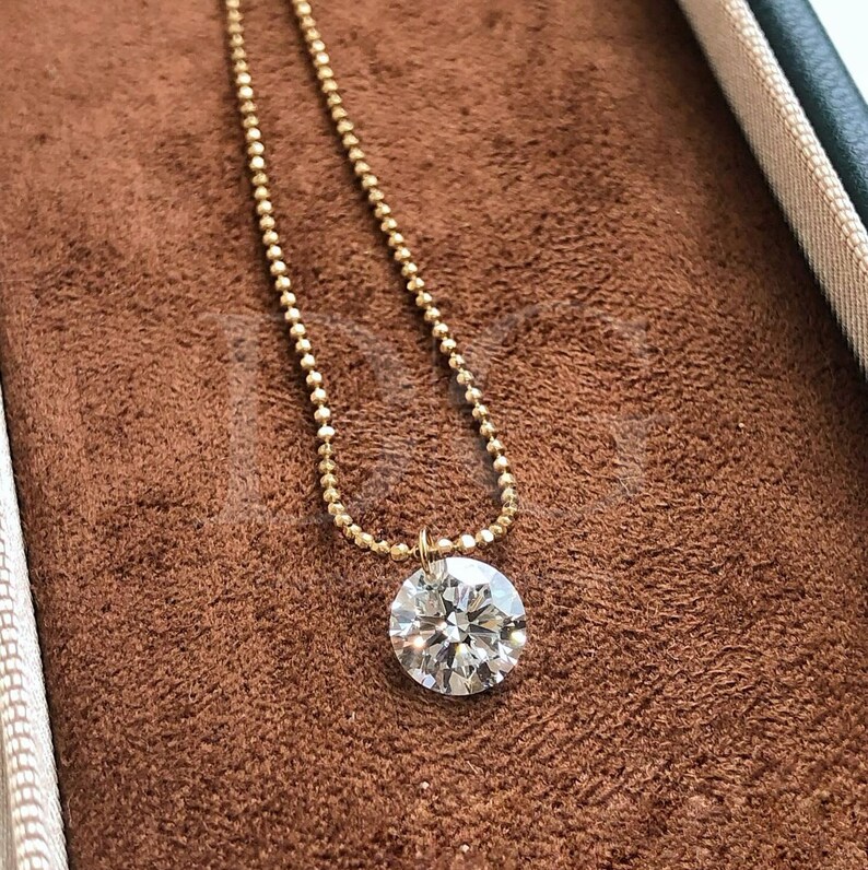 May include: A gold necklace with a round, clear diamond pendant. The diamond is set in a simple bezel and hangs from a delicate beaded chain. The necklace is displayed on a brown velvet surface.