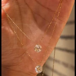 May include: Two delicate gold necklaces, each with a sparkling, round-cut diamond pendant. The necklaces are layered, with one slightly longer than the other. The diamonds catch the light, creating a brilliant display.