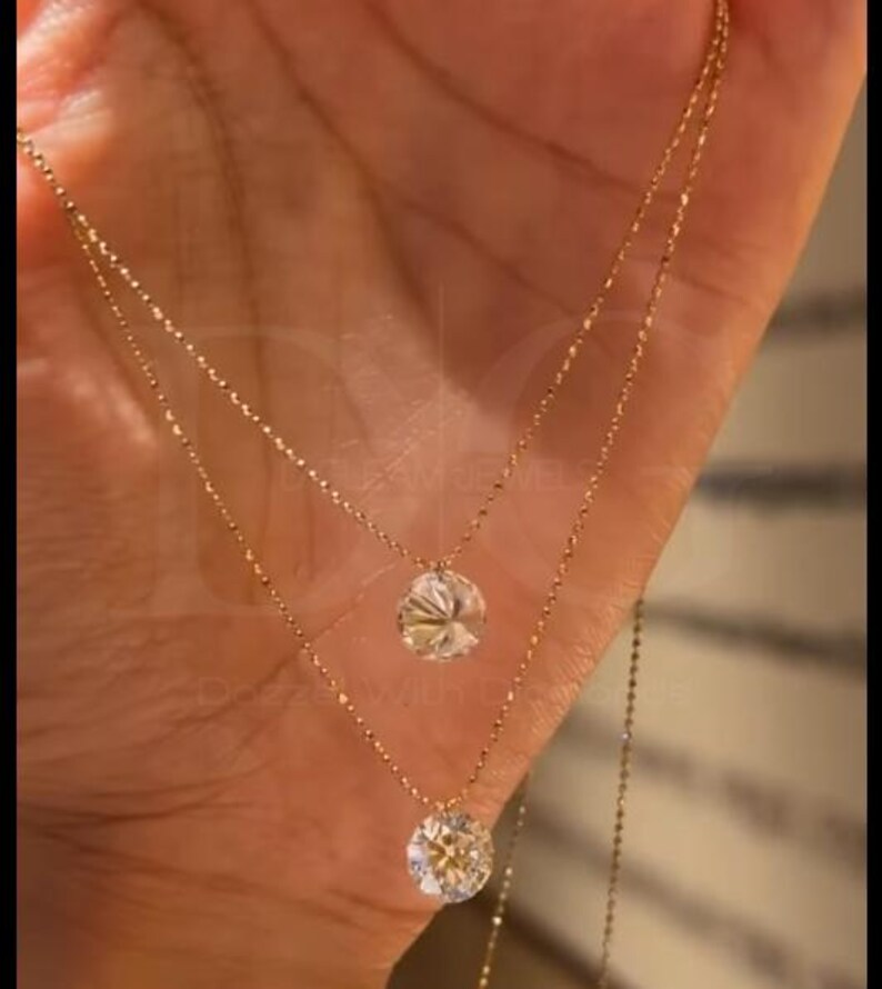 May include: Two delicate gold necklaces, each with a sparkling, round-cut diamond pendant. The necklaces are layered, with one slightly longer than the other. The diamonds catch the light, creating a brilliant display.