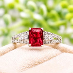 May include: A silver-toned ring featuring a large, rectangular, faceted red gemstone at the center. The band is split into two sections on each side, adorned with small, clear, round-cut stones embedded along the metal surface.
