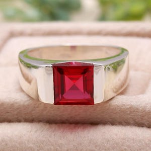 May include: A silver ring with a striking, square-cut, bright red gemstone. The ring has a minimalist design with a shiny finish. The gemstone is the main feature, centrally set within the band.