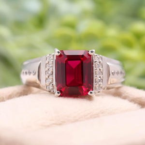 Puede incluir: Un anillo plateado con una gran gema roja oscura rectangular como pieza central. El anillo está flanqueado por gemas claras más pequeñas a cada lado, sobre un fondo suave y neutro.