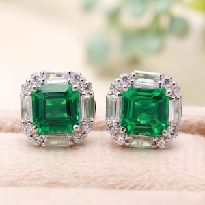 May include: A pair of square-cut emerald earrings with a halo of small, round, and baguette-cut white gemstones. The earrings are set in a silver-colored metal and are displayed on a light pink surface.