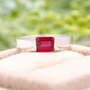 May include: A silver ring featuring a rectangular, vivid red gemstone. The ring has a simple, modern design, highlighting the gemstone's colour. The ring is presented on a light-coloured, textured surface.