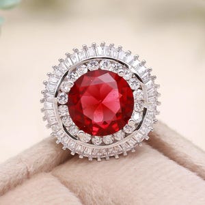 May include: A large, round ring with a vibrant red gemstone center. The gemstone is surrounded by a halo of smaller, clear stones, set in a silver-colored band. The ring is displayed on a soft, beige surface.
