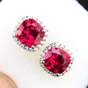 May include: A pair of red gemstone earrings with a halo of smaller white gemstones. The earrings are set in a silver metal.