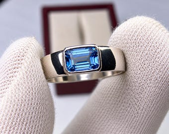 May include: A silver-toned metal ring featuring a single rectangular emerald-cut blue gemstone set in a smooth bezel setting. The band has a polished, reflective surface and a wide, solid construction that tapers slightly toward the center setting.