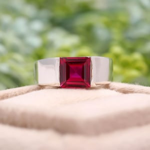 May include: A silver ring featuring a large, square-cut, vibrant red gemstone. The ring has a wide band and a polished finish, showcasing the bold, eye-catching gemstone. The setting is simple, allowing the ruby to be the focal point.