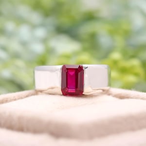 May include: A silver ring featuring a rectangular, deep red gemstone. The ring is displayed on a soft, beige surface, with a blurred green backdrop.