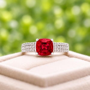 May include: A silver ring featuring a large, cushion-cut, vibrant red gemstone at the center, held by four prongs. The band is wide and encrusted with multiple rows of small, clear, sparkling white stones that extend along the sides.