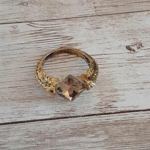 Peut inclure: Une bague dorée ornée d'une pierre précieuse marron clair taillée en carré au centre. L'anneau de la bague présente un motif texturé et complexe sur toute sa surface, s'étendant du sertissage central jusqu'aux côtés de l'anneau circulaire.
