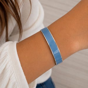 Peut inclure: Un bracelet jonc en émail bleu clair avec une bordure argentée. Le bracelet est porté au poignet, mettant en valeur sa surface lisse et brillante. Un bijou simple et élégant, parfait pour accessoiriser.