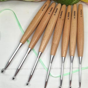 May include: A collection of seven crochet hooks, each with a wooden handle and a silver metal hook. Sizes are marked from 3.5mm to 6.5mm. The light brown wooden handles contrast with the silver hooks.