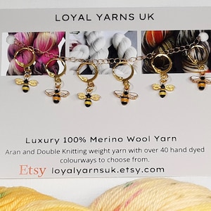 May include: A set of six gold-tone stitch markers, each with a bee charm in black and yellow. The markers are linked by a chain. The card behind them reads "LOYAL YARNS UK" and "Luxury 100% Merino Wool Yarn". The text also mentions hand-dyed yarn.