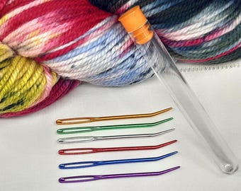 May include: A colorful skein of yarn with a clear tube and six curved, colorful yarn needles. The yarn is a mix of pink, yellow, blue, and green. The needles are gold, green, silver, red, blue, and purple.