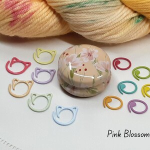 May include: A collection of colorful knitting stitch markers and a floral-decorated container. The stitch markers are in various colors, including pink, yellow, blue, and green. The text "Pink Blossom" is at the bottom.