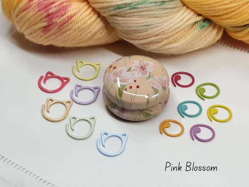 May include: A collection of colorful knitting stitch markers and a floral-decorated container. The stitch markers are in various colors, including pink, yellow, blue, and green. The text "Pink Blossom" is at the bottom.