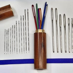 May include: A wooden needle case with a gold band contains various needles in different sizes and colours. The needles are arranged on a white surface with a blue ribbon. The case is open, displaying the needles.