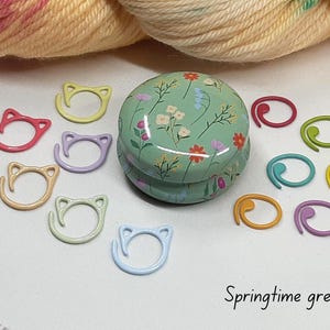May include: A collection of colorful knitting stitch markers and a floral-patterned tin. The stitch markers are in various colors, including pink, yellow, and blue, and are shaped like cat faces. The tin is green with a floral design.