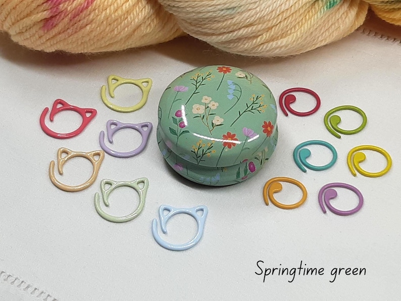 May include: A collection of colorful knitting stitch markers and a floral-patterned tin. The stitch markers are in various colors, including pink, yellow, and blue, and are shaped like cat faces. The tin is green with a floral design.