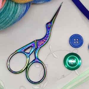 May include: Iridescent stork scissors, with a colourful finish, next to blue buttons, a green button, and colourful yarn. A blue ribbon and a green ribbon are also present. The scissors are designed to resemble a stork.