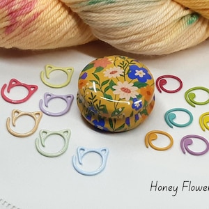 May include: A collection of colorful knitting stitch markers and a floral-patterned tin. The stitch markers are in various colors, including pink, yellow, and blue. The tin is yellow with a floral design. The text "Honey Flower" is visible.