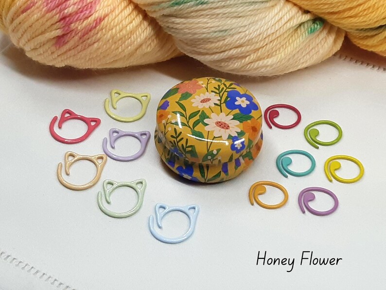 May include: A collection of colorful knitting stitch markers and a floral-patterned tin. The stitch markers are in various colors, including pink, yellow, and blue. The tin is yellow with a floral design. The text "Honey Flower" is visible.