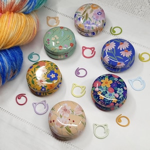 May include: Several small, round tins with floral designs in shades of blue, yellow, and pink are displayed on a white cloth.  Yarn and stitch markers in various colours and shapes are also present, suggesting a crafting theme.