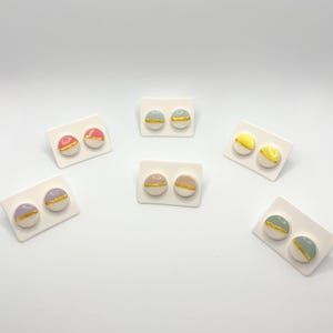 May include: Six pairs of ceramic stud earrings with gold accents. Each pair is displayed on a white card. The earrings are in various colors, including pink, purple, yellow, and blue.