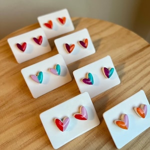 Può includere: Sei paia di orecchini a forma di cuore su carte bianche. Gli orecchini sono in ceramica e sono dipinti in vari colori, tra cui rosso, rosa, arancione, verde acqua e viola.