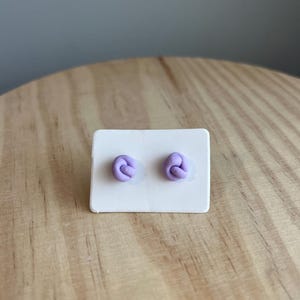 Könnte beinhalten: Ein Paar violette Polymer Clay Ohrringe in Knotenform. Die Ohrringe werden auf einer weißen Karte präsentiert.