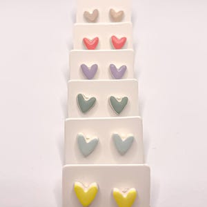 Puede incluir: Un conjunto de pendientes en forma de corazón de varios colores, incluyendo amarillo, azul, verde, morado y rosa. Los pendientes se muestran en una tarjeta blanca.