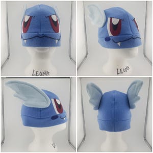 May include: Blue fleece hat with a dragon face design. The hat has white ears and red eyes. The hat is on a white mannequin head. The text "LEONA" is written on the base of the mannequin.