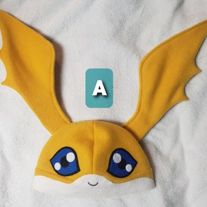 May include: A yellow and white plush hat with large ears shaped like wings. The hat has blue eyes with white pupils and a smiling mouth.
