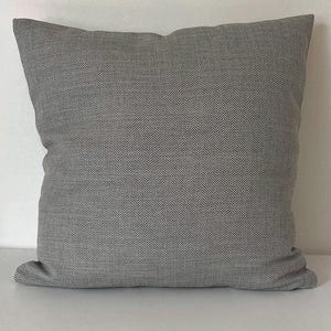 May include: A square throw cushion featuring a grey and white herringbone pattern. The fabric has a woven texture with a consistent repeating V-shaped design across the entire surface of the cushion, creating a neutral appearance.