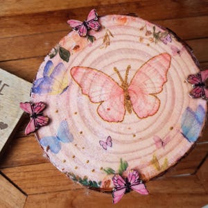Może przedstawiać: Okrągły plaster drewna ozdobiony dużym różowym motylem i mniejszymi motylami. Plaster drewna ma jasnoróżowe tło z kwiatowymi i motylkowymi akcentami. Do plastra drewna przymocowano kilka różowych ozdób w kształcie motyli.