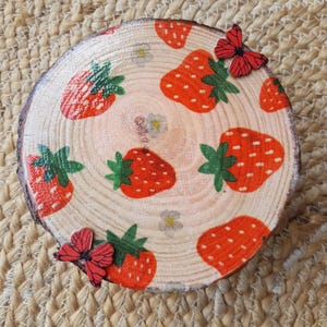 May include: A circular wooden coaster decorated with a strawberry pattern. The coaster displays red strawberries with green leaves and small white flowers. Two red butterfly accents are placed on the coaster.